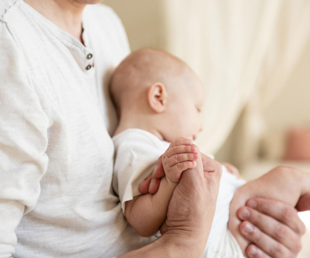 Osoba trzymająca niemowlę w ramionach. O pielęgnacji skóry głowy przy ciemieniusze przeczytasz na MJM.