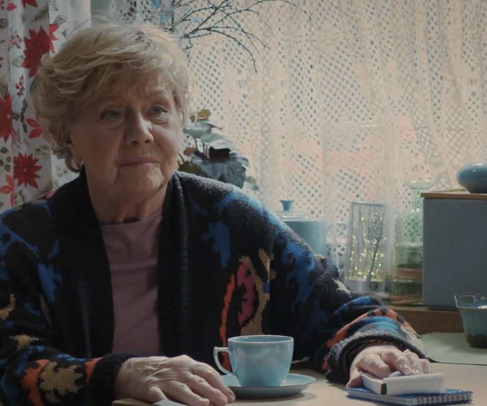 Barbara Mostowiakowa (Teresa Lipowska) siedząca przy stole, na którym leży jasnoniebieska filiżanka i spoczywają jej dłonie. Ma na sobie kolorowy sweter z wzorami w odcieniach czerni, błękitu i pomarańczu. Za nią widać wzorzystą zasłonę i białą koronkową firankę. Ten kadr z M jak miłość ilustruje intuicję Mostowiakowej, o której przeczytasz na Super Seriale.