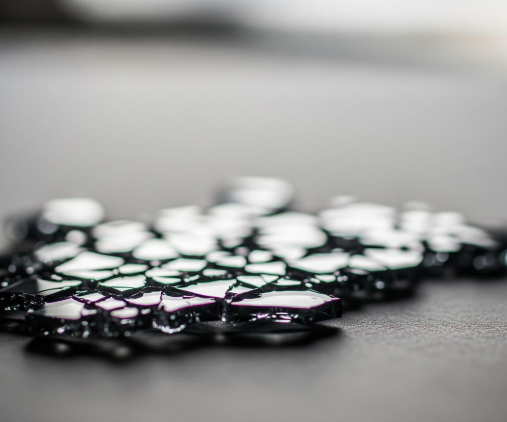 Na ciemnoszarym, teksturowanym tle rozsypane są liczne drobne, błyszczące kawałki o nieregularnych kształtach, przypominające rozbite szkło lub lód. Fragmenty te, o lekko fioletowym zabarwieniu na krawędziach, intensywnie odbijają światło, tworząc białe plamy i rozświetlenia na ich górnych powierzchniach. Tło za rozsypanymi kawałkami staje się coraz jaśniejsze i bardziej rozmyte w kierunku góry obrazu, przechodząc w niemal białą poświatę.