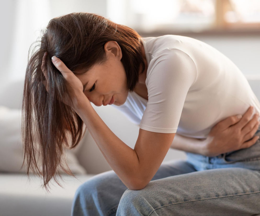 Kobieta siedząca na kanapie z dłonią przy skroni i drugą dłonią na brzuchu, cierpiąca na ból. Zdjęcie ilustruje dolegliwości trawienne i potrzebę domowych sposobów na ból brzucha, o których można przeczytać na portalu Poradnik Zdrowie.