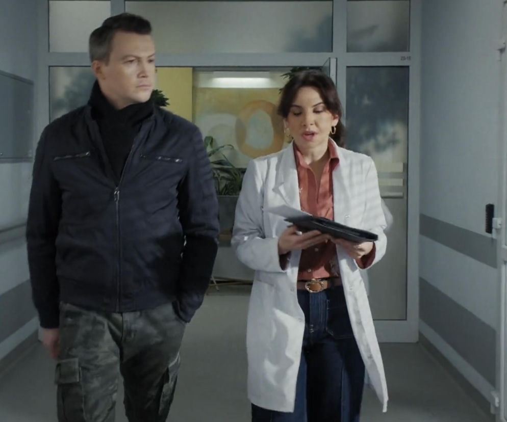 Aktor Krzysztof Kwiatkowski jako Jakub w ciemnej kurtce i Ilona Janyst jako Aneta w białym lekarskim kitlu, idą korytarzem kliniki, Aneta trzyma dokumenty i patrzy na nie, Jakub spogląda w bok. Scena z serialu M jak miłość. Więcej o serialu przeczytasz na Super Seriale.