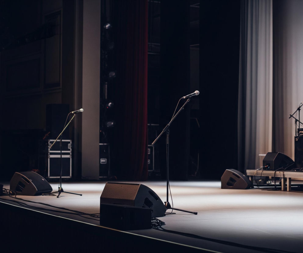 Scena z ustawionym sprzętem muzycznym i oświetleniem, gotowa do występu. Na pierwszym planie, po lewej stronie, znajdują się dwa monitory sceniczne oraz stojak z mikrofonem. Dalej w głąb sceny widoczne są kolejne stojaki mikrofonowe, a także wzmacniacze, głośniki i czarne skrzynie transportowe. Tło sceny tworzy ciemna ściana z fragmentami czerwonej i białej kurtyny po prawej stronie, a wszystko oświetlone jest punktowym światłem padającym na środek sceny.
