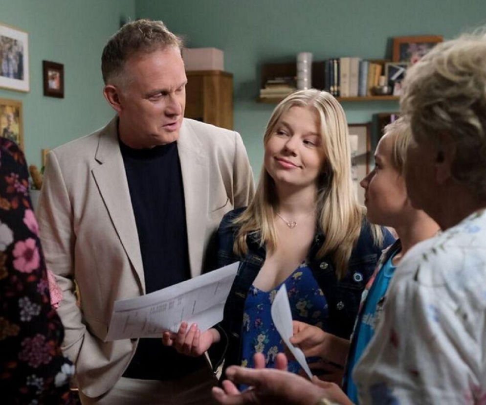 Grupa czterech osób w pomieszczeniu, z mężczyzną w beżowej marynarce i młodą blondynką w jeansowej kurtce, którzy trzymają dokumenty. Konwersacja, kontekst artykułu sugeruje pożegnanie z serialem M jak miłość. Więcej o tym przeczytasz na Super Seriale.