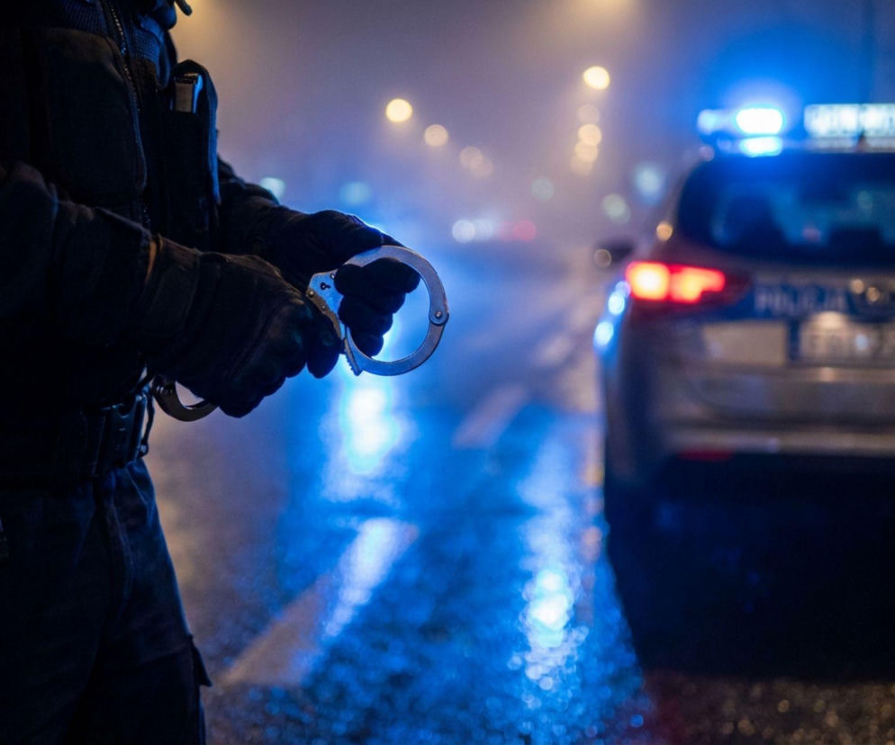 Dłonie policjanta trzymające kajdanki na tle radiowozu