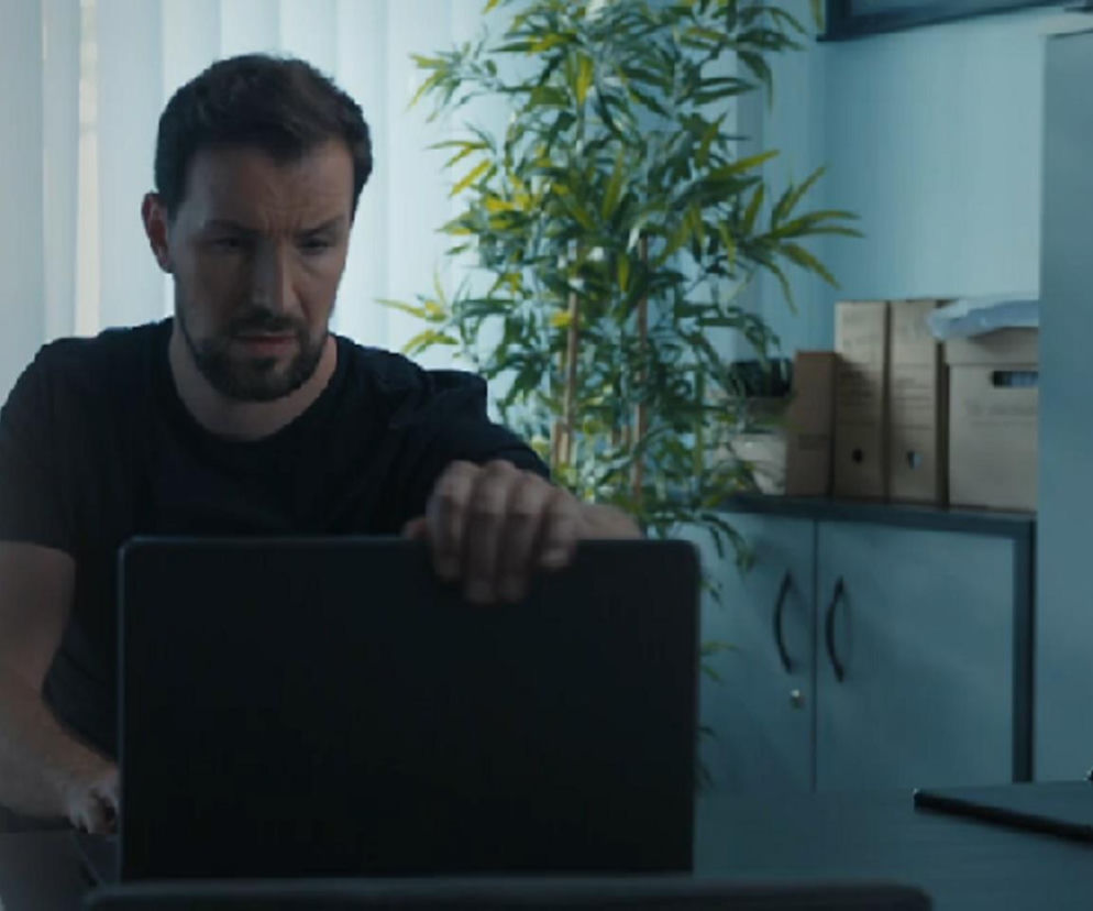 Oskar Stoczyński jako Marcin Kodur, skupiony mężczyzna z brodą w czarnej koszulce, otwiera laptopa w biurze. Za nim widać roślinę i szafę z dokumentami. Scena z serialu Barwy szczęścia o przełomie w śledztwie. Więcej o odcinku 3272 na Super Seriale.
