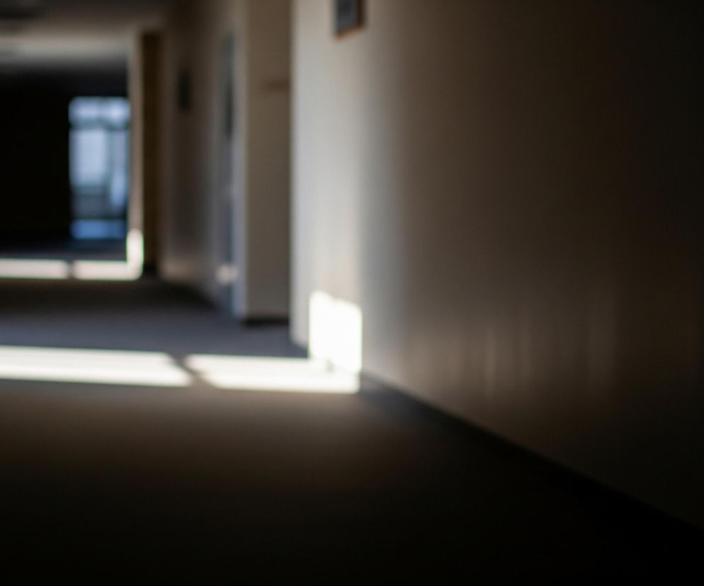 Wąski, rozmyty korytarz dominuje na zdjęciu, oświetlony naturalnym światłem słonecznym tworzącym jasne plamy na podłodze i ścianach. Po lewej stronie, w głębi, widać jaśniejsze pomieszczenie z niewyraźnymi elementami okna, a wzdłuż korytarza rozciągają się ciemne, poziome i pionowe cienie. Prawa ściana jest jaśniejsza u góry, przechodząc w ciemny brązowy odcień ku dołowi, z widocznym podłużnym blaskiem światła w dolnej części.