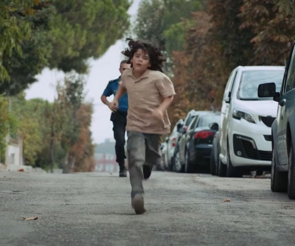 Mały Yusuf z tureckiego serialu Dziedzictwo biegnący ulicą w beżowej koszulce i ciemnych spodniach. Za nim widać biegnącą osobę. Obok zaparkowane są samochody, a w tle zieleń. Widzowie Super Seriale z niepokojem śledzą losy zagubionego chłopca.