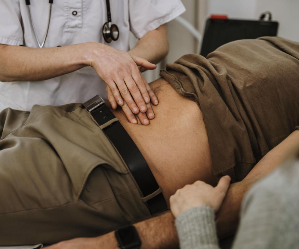 Lekarz z stetoskopem wykonuje badanie palpacyjne brzucha pacjenta leżącego na kozetce. To kluczowy etap w diagnostyce chorób jelit, o których możesz przeczytać więcej na Poradnik Zdrowie.