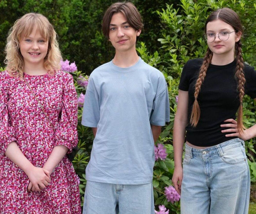 Trójka nastolatków, Emilka (Jessica Frankiewicz) w kwiecistej sukience, Emil (Artem Malaszczuk) w błękitnym T-shircie i Pola (Gabriela Szewczyk) w okularach z warkoczami, uśmiecha się na tle kwitnących rododendronów. Fotografia promuje rywalizację o serce Emila, o której przeczytasz na Super Seriale.