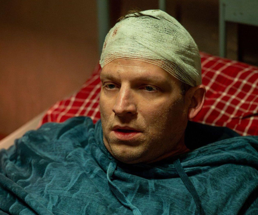 Mężczyzna z opatrunkiem na głowie, ubrany w niebieską bluzę, leży na łóżku z czerwoną pościelą w kratę, obudzony w piwnicy. Jest to Łukasz (Michał Rolnicki) z serialu Barwy szczęścia, porwany i uwięziony, o czym przeczytasz na Super Seriale.