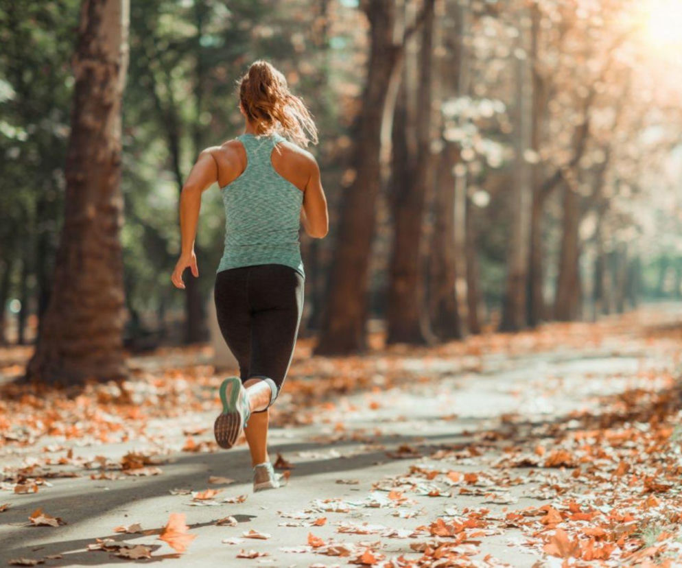 Kobieta biegnąca po ścieżce w lesie, otoczona drzewami i spadającymi liśćmi, symbolizuje aktywność fizyczną i profilaktykę kontuzji. Na Poradnik Zdrowie znajdziesz wskazówki, jak unikać urazów podczas biegania.