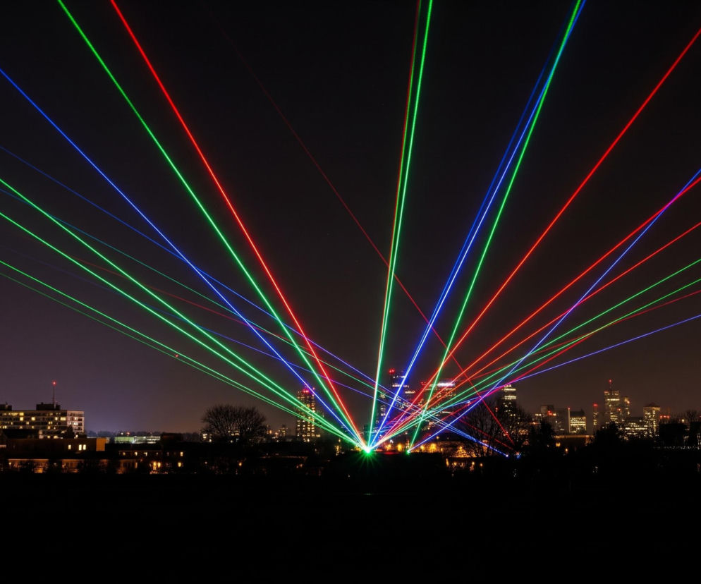 W centrum kadru widoczne są liczne, jaskrawe promienie laserowe w kolorach zielonym, czerwonym i niebieskim, rozchodzące się wachlarzowo z jednego punktu na dole pośrodku. Promienie te przecinają ciemne, nocne niebo nad panoramą miasta. W tle, pod smugami światła, rozciąga się miejski krajobraz z licznymi, rozświetlonymi budynkami, z których niektóre to wieżowce z czerwoną lampą na szczycie. Na pierwszym planie, w dolnej części obrazu, widoczna jest ciemna sylwetka terenu, prawdopodobnie z drzewami i niską roślinnością, z nielicznymi, rozproszonymi światłami.