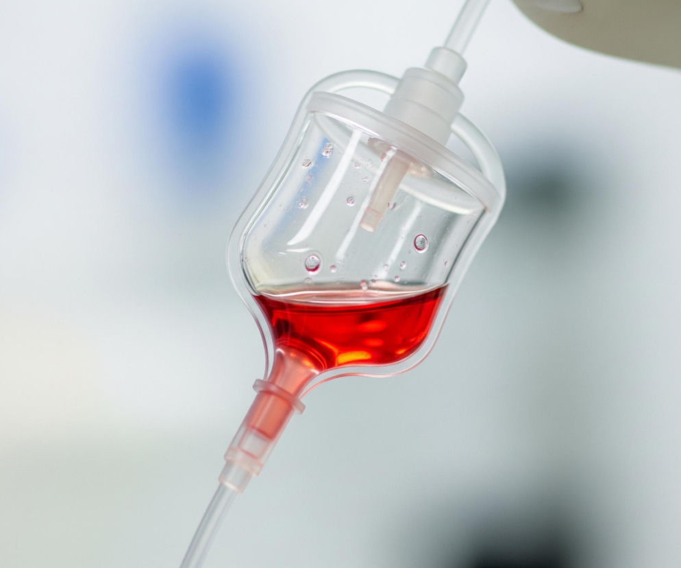 Przezroczysta komora kroplowa, wypełniona do połowy intensywnie czerwoną cieczą, zawieszona jest na tle jasnego, rozmytego tła. Z górnej części komory odchodzi krótki, przezroczysty element łączący ją z niewidocznym źródłem, natomiast z dolnej części wychodzi cieńszy, przezroczysty dren prowadzący w dół. Wewnątrz czerwonej cieczy widoczne są drobne pęcherzyki powietrza, a na ściankach górnej, pustej części komory również kilka małych kropel. Tło jest jasnoszare z lekko rozmazanym niebieskim akcentem po lewej stronie.