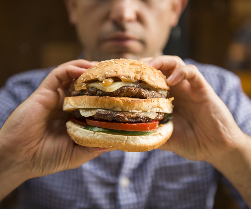 Mężczyzna w koszuli w kratę trzyma w dłoniach dużego hamburgera z podwójnym kotletem, serem, cebulą, pomidorem i ogórkiem. Ten obraz ilustruje temat śmiertelnej alergii na mięso, o której przeczytasz więcej na portalu Poradnik Zdrowie.