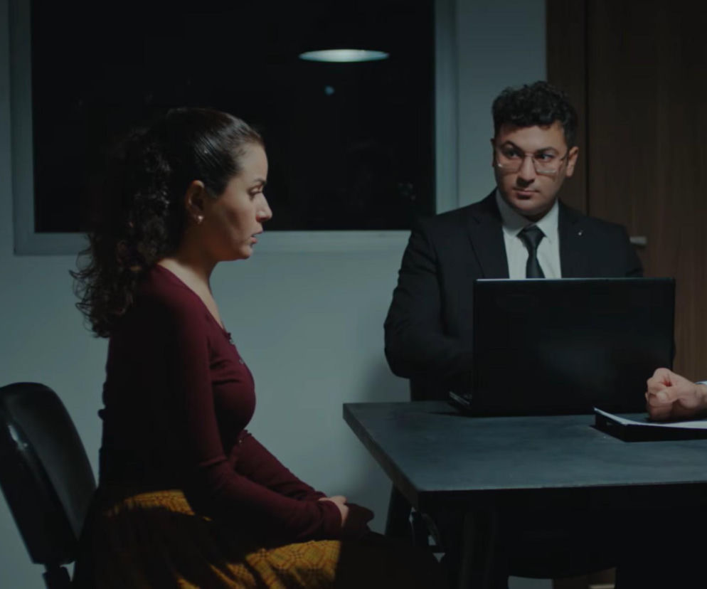 Aynur, kobieta z długimi, ciemnymi włosami związanymi w kucyk, w bordowej bluzce i żółtej spódnicy, siedzi przy ciemnym stole podczas przesłuchania policyjnego, spuszczając wzrok. Naprzeciwko niej siedzi prokurator w garniturze i okularach, z laptopem na stole, a obok widać rękę innej osoby z dokumentami. Dowiedz się więcej o jej kłopotach na naszym portalu.
