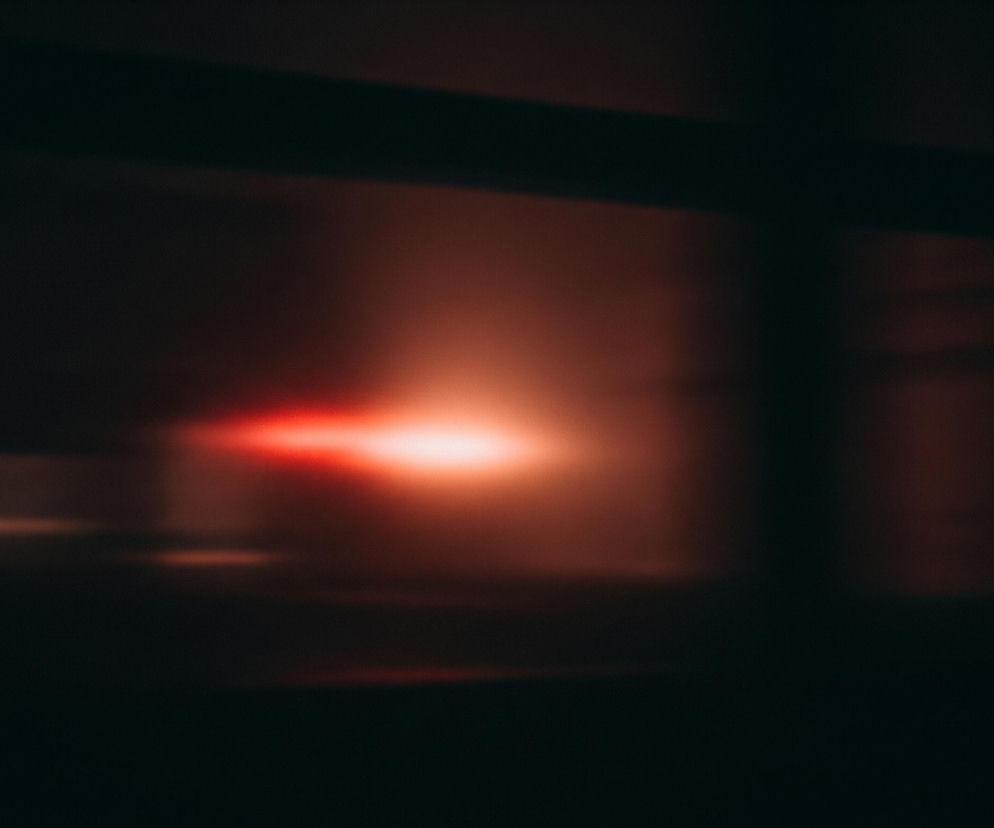 Główny plan to ciemne, rozmyte tło, które stopniowo jaśnieje ku prawej i górnej części, przyjmując odcienie brązu i czerwieni. W centrum kadru znajduje się intensywna, czerwono-pomarańczowa plama światła, rozciągająca się horyzontalnie, z wyraźnym, jaśniejszym punktem po lewej stronie, przypominającym smugę światła. Górna część obrazu ukazuje ciemniejszą, poziomą smugę, która zdaje się przecinać jaśniejszą poświatę tła. Całość jest silnie rozmyta, co nadaje zdjęciu abstrakcyjny charakter.