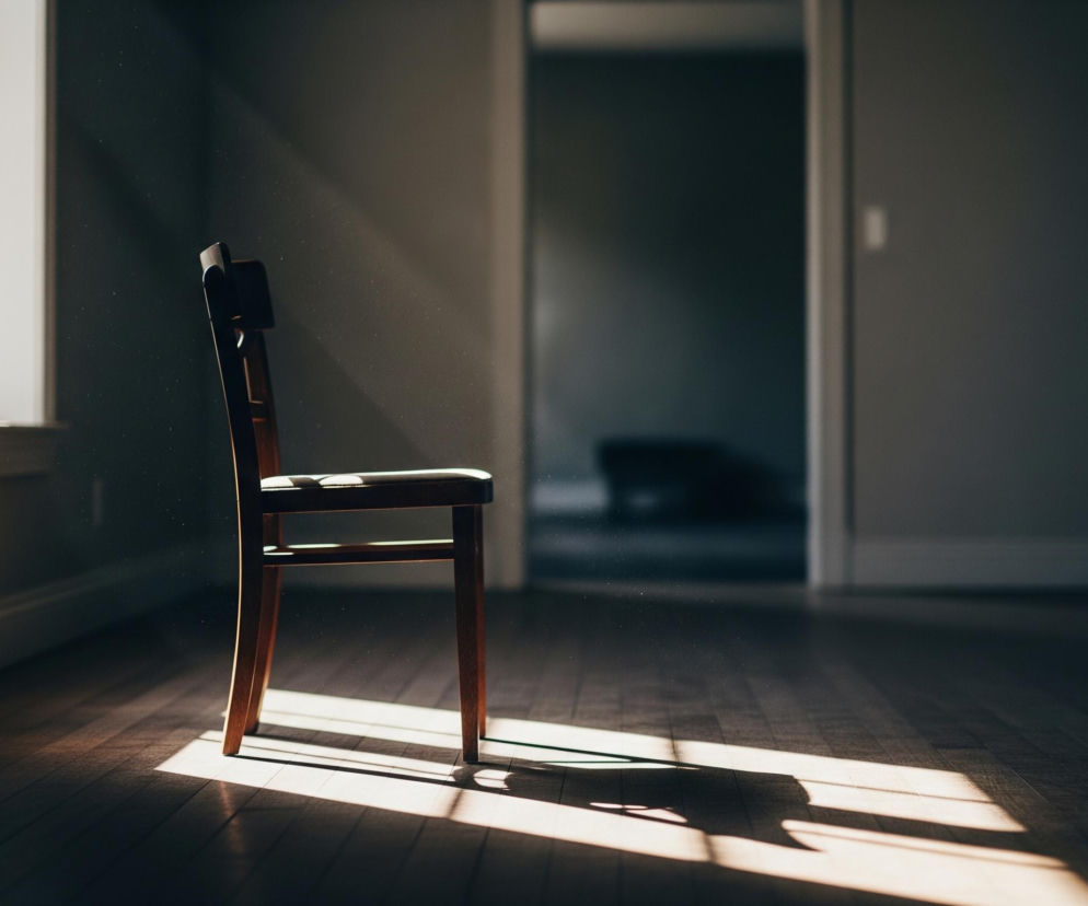 Drewniane krzesło z ciemnym siedziskiem stoi w półmroku, oświetlone promieniami słońca wpadającymi przez okno po lewej stronie. Na podłodze z drewnianych desek widoczne są długie, kontrastowe cienie rzucane przez krzesło i okno. W powietrzu, w snopach światła, unoszą się drobne pyłki, a w tle widać otwarte drzwi prowadzące do innego, ciemniejszego pomieszczenia.