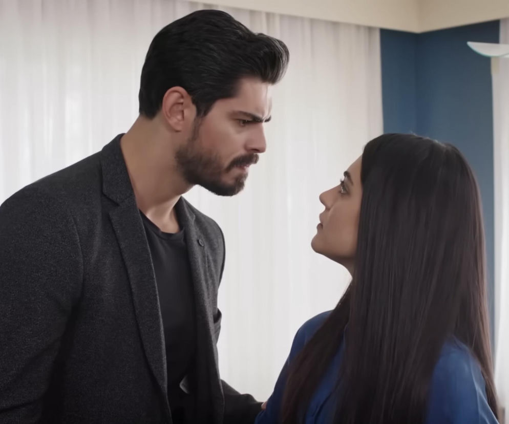 Mężczyzna z brodą, Cihan, i kobieta z długimi włosami, Hancer, patrzą na siebie z napięciem podczas kłótni w sypialni. Hancer ma na sobie niebieską koszulę, Cihan ciemną marynarkę. Scena z odcinka serialu Panna Młoda. Więcej o serialach na Super Seriale.