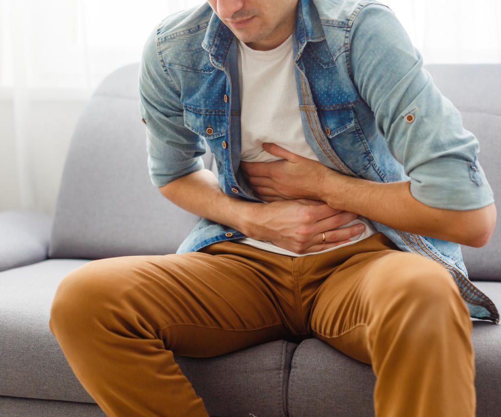 Mężczyzna w jasnej jeansowej koszuli i brązowych spodniach siedzi na szarej kanapie, trzymając się za brzuch, co symbolizuje silny ból brzucha i problemy trawienne, typowe dla przewlekłych chorób jelit. Więcej na ten temat przeczytasz na portalu Poradnik Zdrowie.