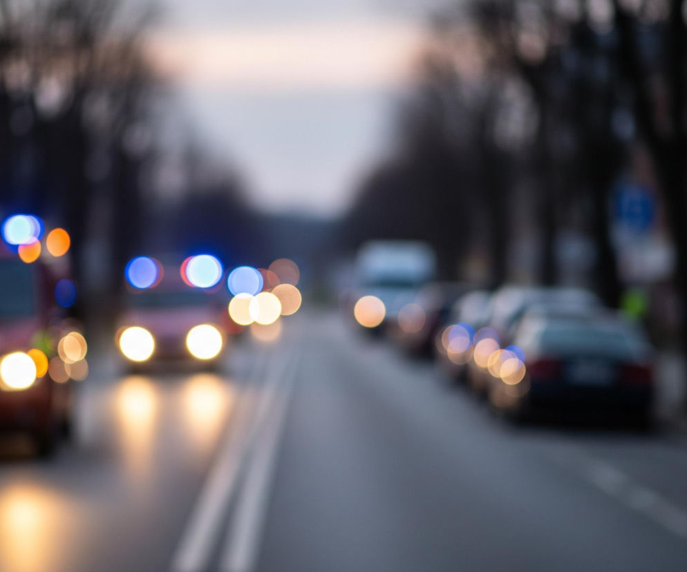 Czerwony pojazd z niebieskimi i pomarańczowymi światłami sygnalizacyjnymi na dachu oraz jasnymi reflektorami zbliża się lewym pasem drogi, z perspektywy fotografującego. Droga rozdziela się na dwa pasy wyraźnymi białymi liniami, które zbiegają się w oddali. Po prawej stronie drogi stoją zaparkowane samochody, a ich światła, wraz z światłami innych pojazdów nadjeżdżających z naprzeciwka, tworzą rozmyte, kolorowe kręgi bokeh w odcieniach pomarańczowego i niebieskiego. Tło stanowią rozmyte kształty drzew i budynków, a w górnej części obrazu widoczne jest jasne, różowo-niebieskie niebo o zmierzchu.