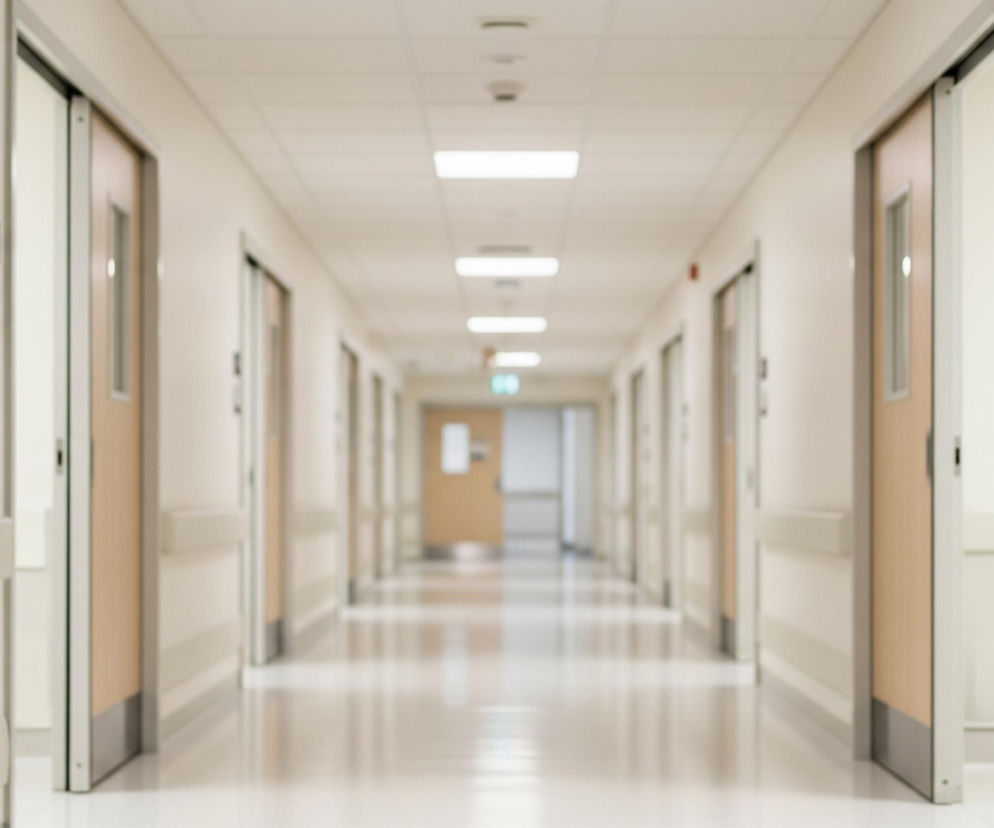 Długi, symetryczny korytarz szpitalny rozciąga się w głąb obrazu, zbiegając się w odległym punkcie. Po obu stronach korytarza widoczne są równomiernie rozmieszczone drzwi, a ich jasnobrązowe panele i ciemniejsze futryny kontrastują z kremowymi ścianami. Na ścianach, na wysokości około metra, biegną poziome, jasnoszare listwy ochronne. Błyszcząca, jasna podłoga odbija światło z sufitu, gdzie umieszczono szereg prostokątnych opraw oświetleniowych, rozmieszczonych wzdłuż centralnej osi korytarza.