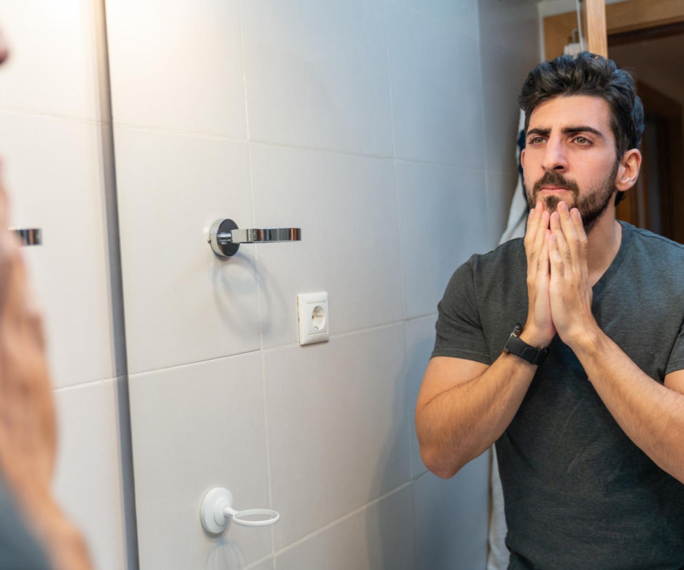 Młody mężczyzna z brodą i ciemnymi włosami w szarej koszulce stoi przed lustrem w łazience, trzymając ręce złączone pod brodą, w zamyśleniu. Prawdopodobnie obserwuje zmiany w swoim ciele, co nawiązuje do tematyki dreszczy w toalecie, o czym można przeczytać na portalu Poradnik Zdrowie.