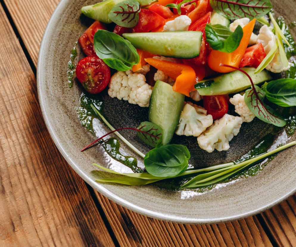Kolorowa sałatka na talerzu z kawałkami pomidorów, ogórków, papryki i kalafiora, polana zielonym sosem pesto, z listkami bazylii i szczypiorkiem, na tle drewnianego stołu. Zdjęcie ilustruje zdrowe odżywianie, o którym przeczytasz więcej na Poradnik Zdrowie.