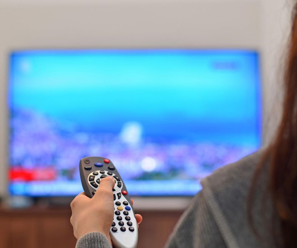 Kobieta trzymająca pilota do telewizora, ustawiająca głośniej kanał na ekranie. Scena symbolizuje pierwsze sygnały problemów ze słuchem, które mogą być powiązane z demencją. Dowiedz się więcej o profilaktyce niedosłuchu na Poradnik Zdrowie.