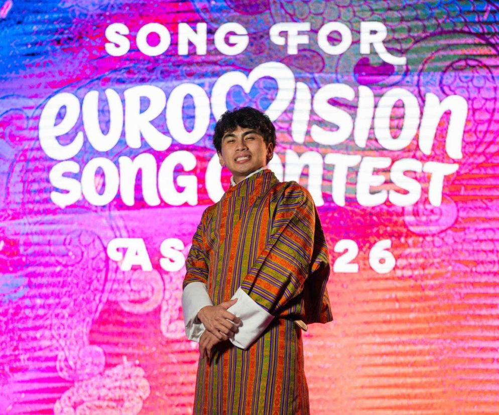 Piosenkarz Nyendra w kolorowym stroju na tle logo Eurowizja Azja 2026. O reprezentancie Bhutanu przeczytasz w Eska.