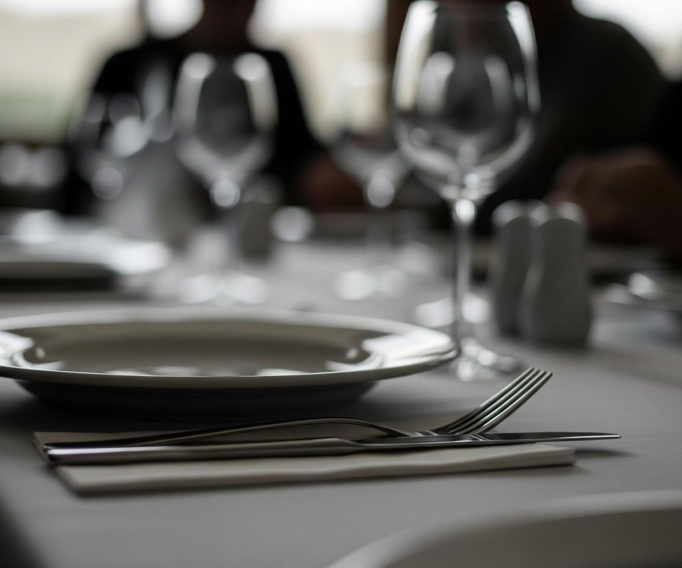 Ustawiony stół restauracyjny z jasnoszarym talerzem na pierwszym planie po lewej stronie. Na białej serwetce obok talerza leżą srebrne sztućce: nóż i widelec. W tle, częściowo niewyraźne, widoczne są inne nakrycia, składające się z kieliszków do wina i małych solniczek lub pieprzniczek, oraz zarysy ludzi siedzących przy stole. Całość oświetlona jest naturalnym, rozproszonym światłem, tworzącym miękkie cienie i odbicia.