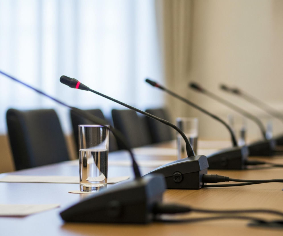 Na drewnianym stole konferencyjnym stoi rząd mikrofonów na wysięgnikach, skierowanych w stronę widza, z jednym szklanką wody pośrodku. Mikrofony są czarne, z subtelnymi czerwonymi akcentami na główkach, a za nimi widać czarne, skórzane oparcia krzeseł. Tło stanowi rozmyta biała ściana z widocznymi, pionowymi fałdami, prawdopodobnie zasłon.