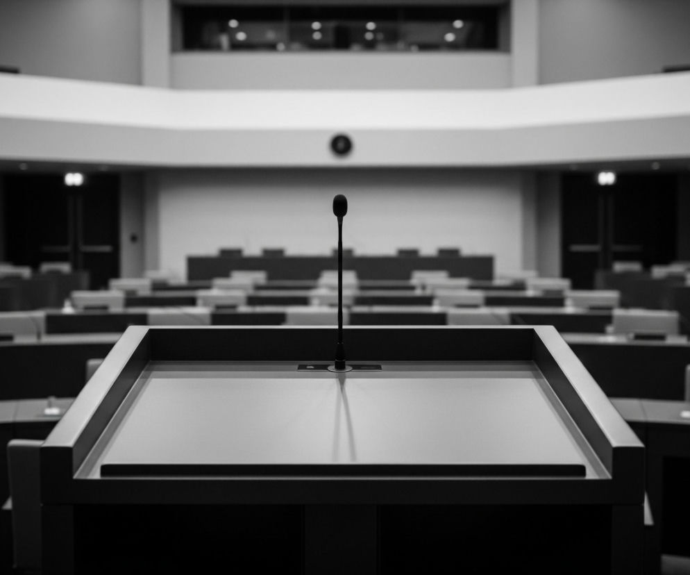 Czarny mikrofon stoi na szarej mównicy w centrum sali konferencyjnej. Sala jest pusta, wypełniona rzędami stołów z ciemnymi krzesłami, skierowanymi w stronę mównicy. Za mównicą, po lewej stronie, widać ciemne, prostokątne otwory w ścianie, a po prawej okrągły, czarny obiekt na ścianie. Dalsze rzędy stołów i krzeseł rozciągają się w głąb sali, a nad nimi, w tle, widoczny jest balkon z niewyraźnymi elementami. Całe pomieszczenie jest utrzymane w odcieniach szarości i czerni, z wyraźnymi kontrastami światła i cienia.