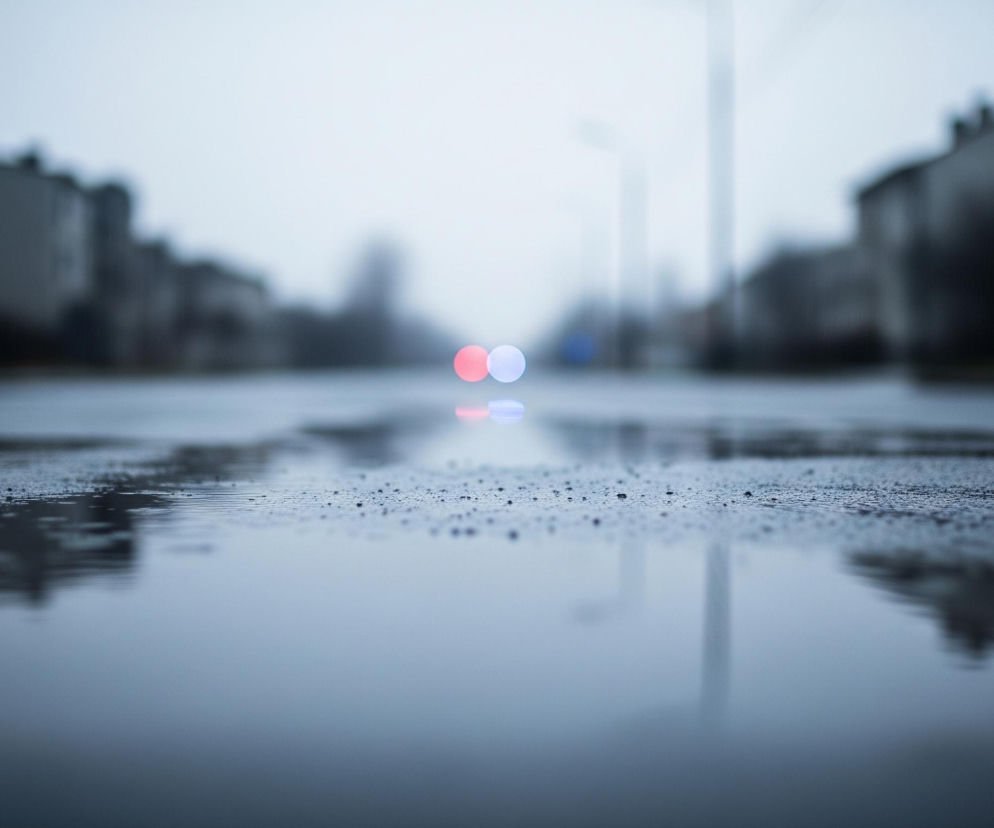 Na pierwszym planie widoczna jest mokra powierzchnia drogi lub chodnika, pokryta licznymi kroplami wody i kałużami, które odbijają otoczenie. W oddali, po obu stronach centralnego punktu, zamazane kontury budynków mieszkalnych i latarni ulicznych rysują się na tle jasnego, zachmurzonego nieba. Na horyzoncie, w centrum kadru, wyróżniają się dwa rozmyte, okrągłe światła: jedno czerwone po lewej i jedno niebieskie po prawej, prawdopodobnie pochodzące od pojazdu, ich odbicia są również widoczne w kałużach. Całość utrzymana jest w chłodnej palecie barw, dominują szarości i błękity, co nadaje obrazowi melancholijny, deszczowy nastrój.