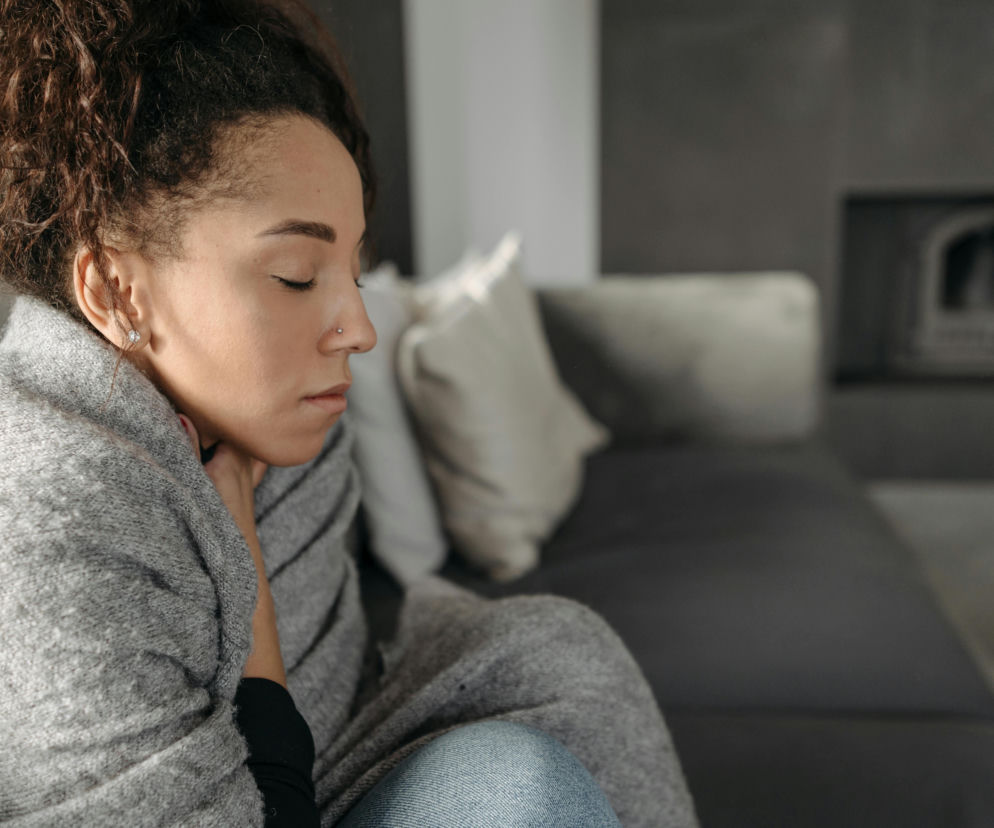 Kobieta z kręconymi włosami, otulona szarym kocem, siedzi na kanapie z zamkniętymi oczami i dłonią przy szyi, symbolizując ból gardła. W tle widoczny jest kominek. Na Poradnik Zdrowie znajdziesz sposoby na ukojenie bólu gardła.