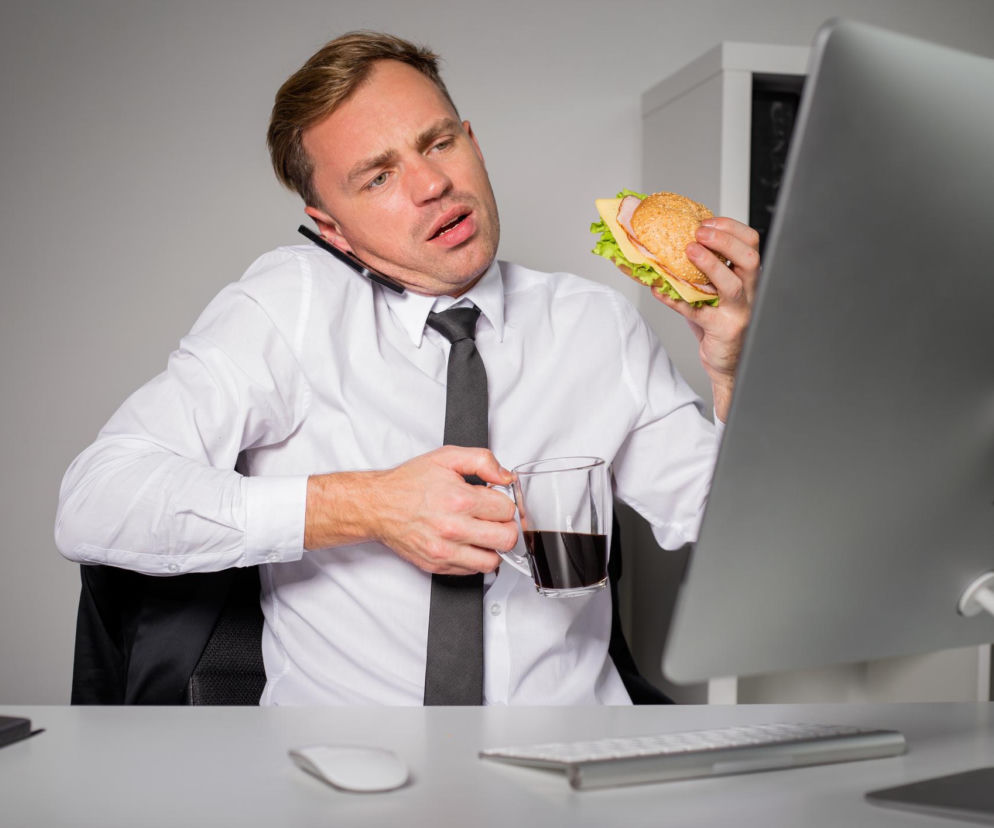 Zapracowany mężczyzna w białej koszuli i krawacie je kanapkę, pije kawę i rozmawia przez telefon, patrząc na ekran komputera, co ilustruje amnezję żywieniową u Polaków. Dowiedz się, jak uniknąć tego problemu, czytając Poradnik Zdrowie.