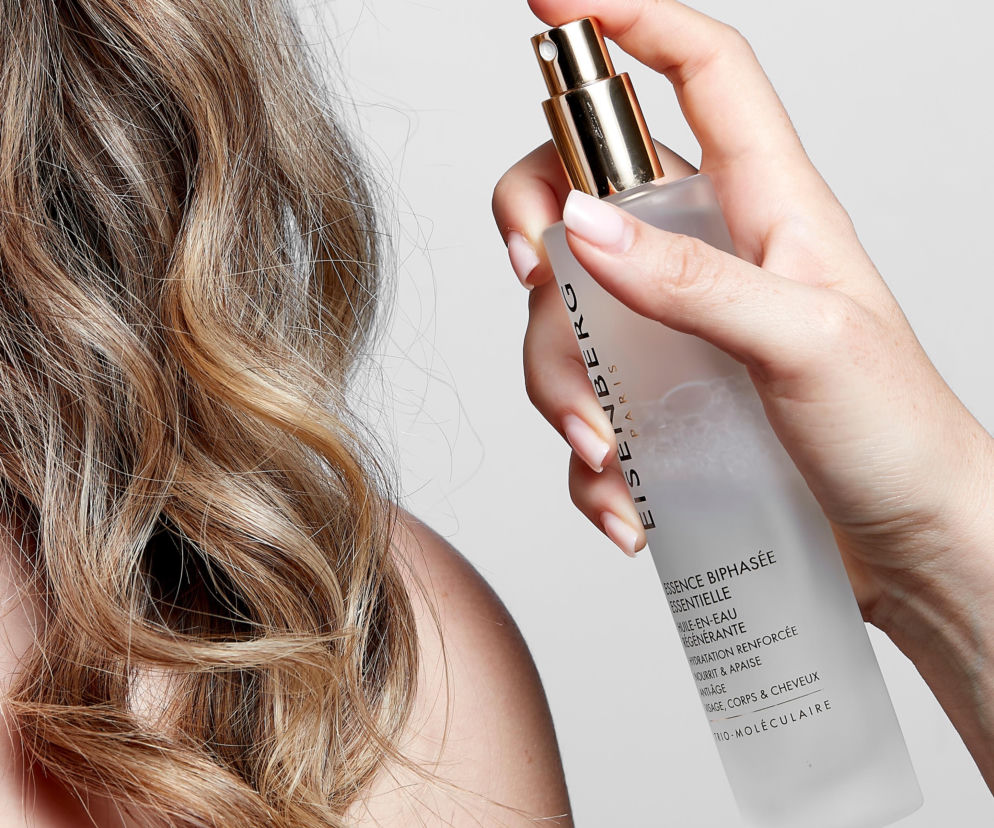 Dłoń kobiety w pastelowym manicure aplikująca Eisenberg Essential Biphase Essence na włosy, widoczne są falowane blond pasma oraz ramię. Ten innowacyjny eliksir regenerujący to produkt 3 w 1, więcej o nim możesz przeczytać na naszym portalu.