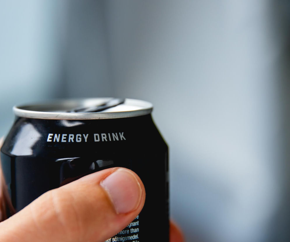 Dłoń trzymająca czarną puszkę napoju energetycznego z białym napisem ENERGY DRINK na pierwszym planie, symbolizującą zagrożenia związane ze spożyciem energetyków. Na portalu Poradnik Zdrowie znajdziesz więcej informacji o ich szkodliwości dla zdrowia.