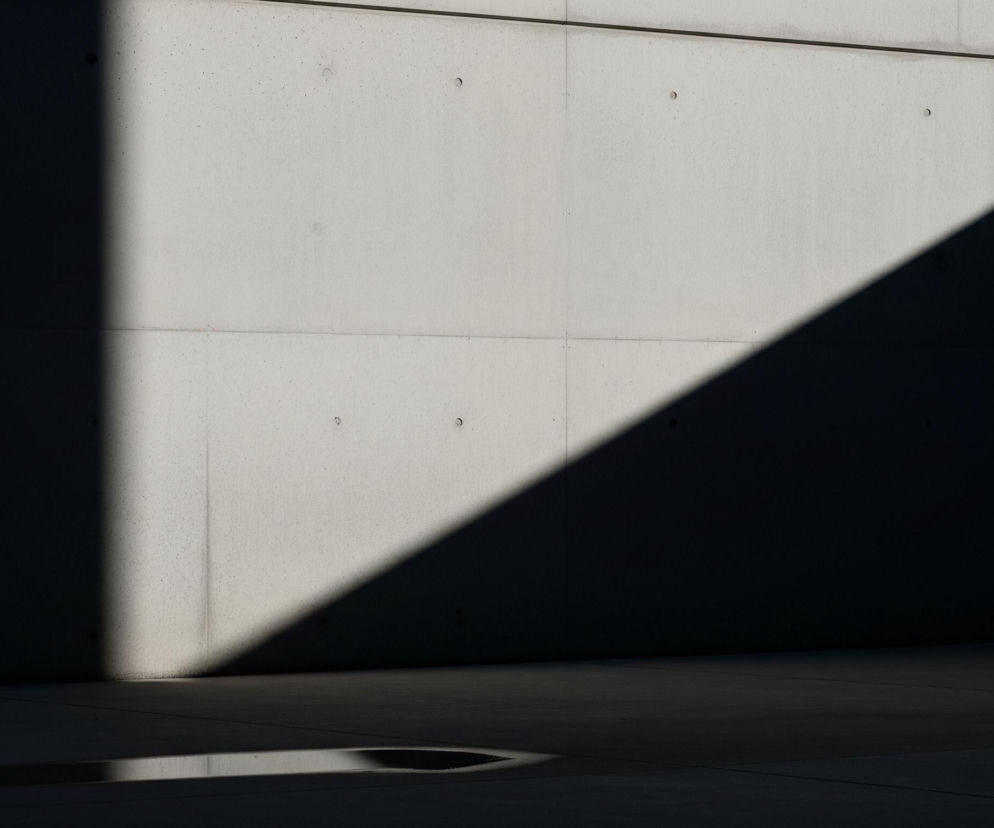 Lewa część obrazu przedstawia jasną, betonową ścianę, oświetloną słońcem, z widocznymi poziomymi i pionowymi liniami podziału oraz kilkoma okrągłymi otworami. Na ścianie i podłodze widoczny jest ostry, skośny cień w kształcie trójkąta, który rozdziela scenę na jasną i ciemną strefę. W prawej, zacienionej części ściany znajduje się otwarte skrzydło drzwi, częściowo odbijające światło, oraz ciemne, puste wnętrze za nimi. Pod otwartymi drzwiami widoczna jest mniejsza, ciemna wnęka.