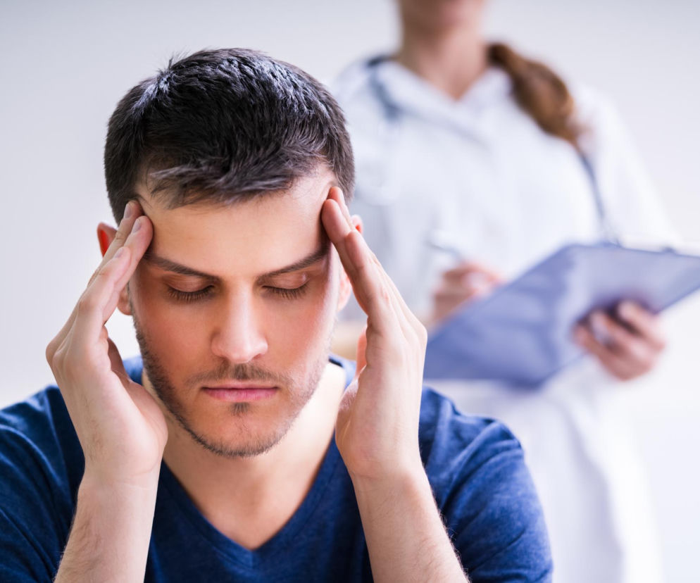 Mężczyzna w niebieskiej koszulce z krótkimi włosami, z zamkniętymi oczami, silnie uciska skronie dłońmi, doświadczając piorunującego bólu głowy. W tle niewyraźna postać lekarki w białym fartuchu, trzymająca podkładkę z dokumentami, symbolizująca szybką diagnostykę, o której przeczytasz na Poradniku Zdrowie.