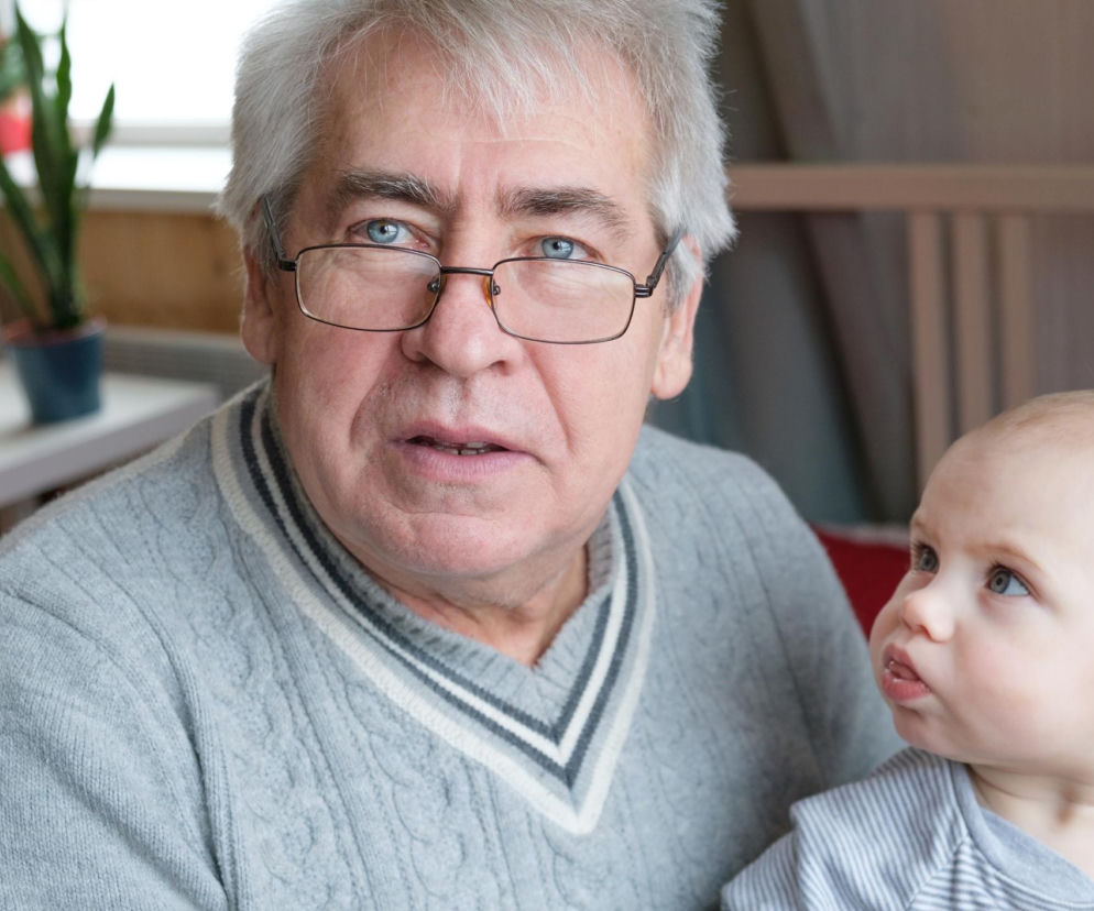 Starszy mężczyzna w okularach i szarym swetrze, z siwymi włosami i niebieskimi oczami, spogląda w obiektyw, trzymając na rękach niemowlę. Niemowlę z jasnymi włosami i niebieskimi oczami patrzy w prawo, w górę. O relacjach z dziadkami, można przeczytać na portalu Poradnik Zdrowie.