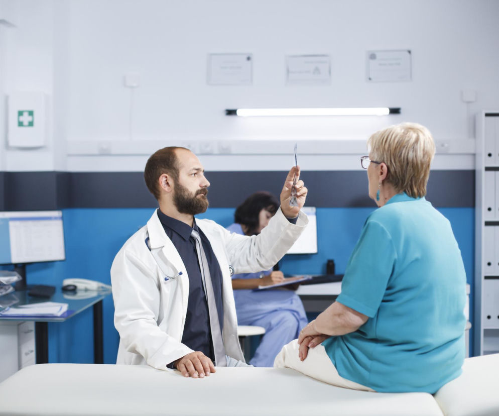 Lekarz z brodą w białym kitlu i stetoskopem na szyi, siedzący na kozetce, pokazuje starszej pacjentce, siedzącej obok, zdjęcie rentgenowskie. Kobieta w turkusowej koszulce ogląda wynik badania. Obraz symbolizuje diagnostykę i leczenie szpiczaka plazmocytowego, o którym przeczytasz na Poradnik Zdrowie.