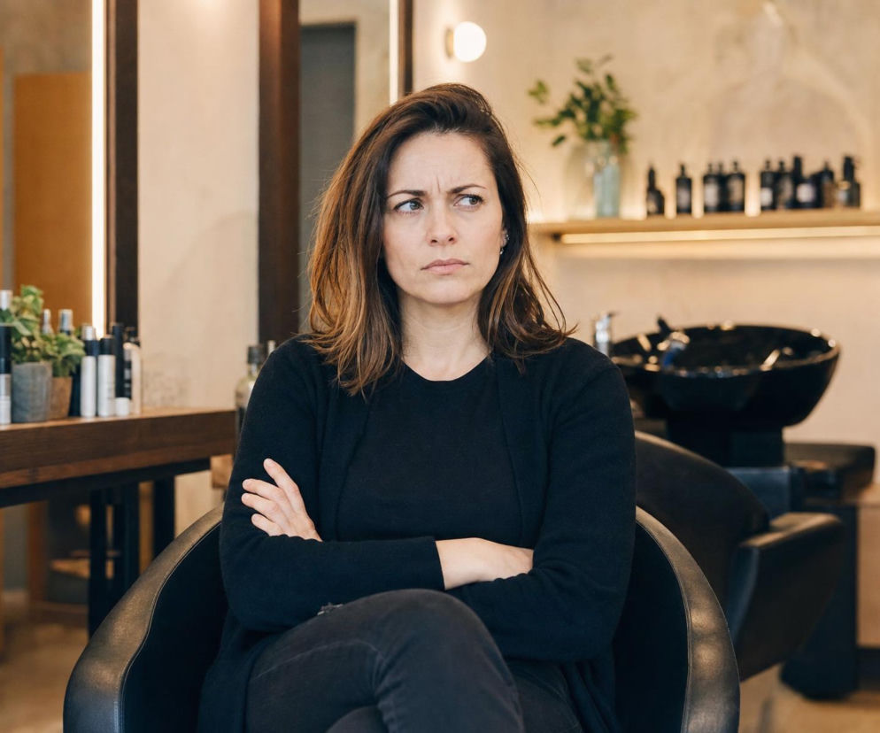 Zasmucona fryzjerka Monika siedzi ze skrzyżowanymi rękami w swoim salonie. Historię złośliwej opinii przeczytasz na SE.