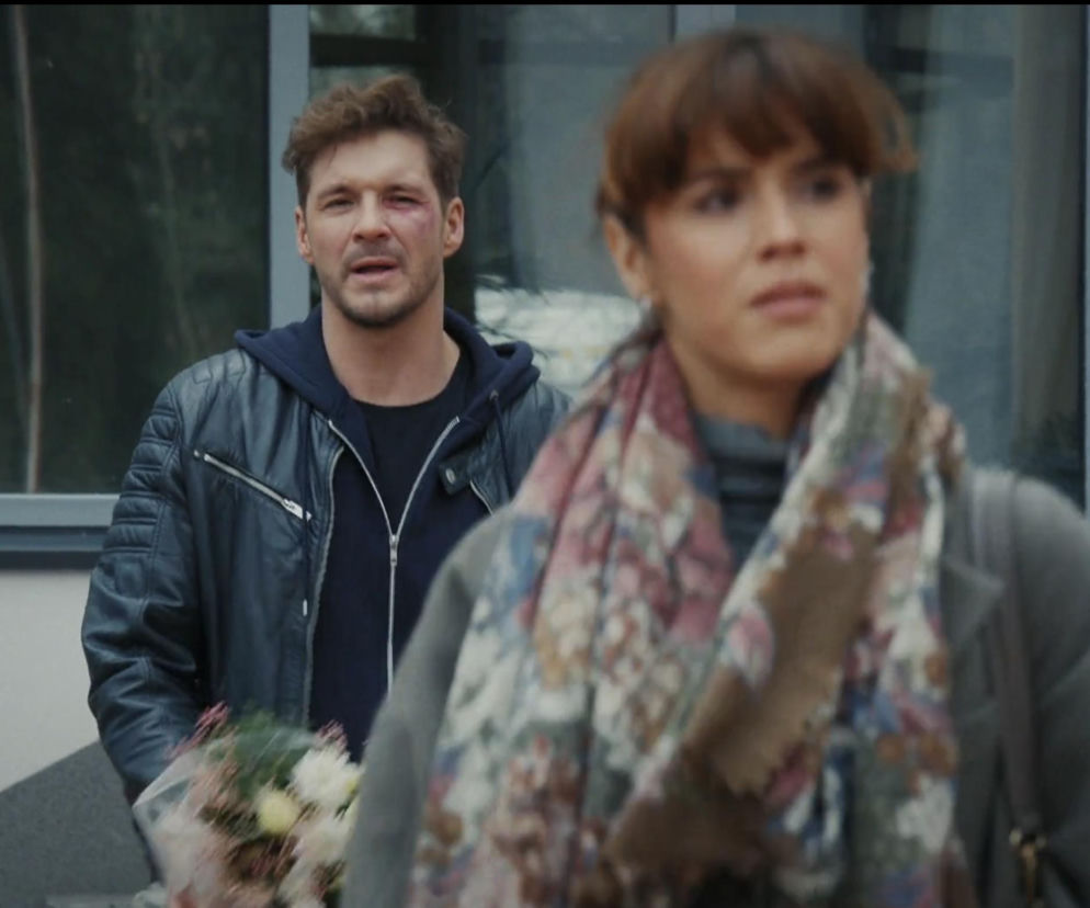 Mężczyzna w czarnej skórzanej kurtce i ciemnej bluzie z kapturem, z widocznym siniakiem pod prawym okiem, trzyma bukiet kwiatów, patrząc na kobietę z przodu w beżowym szaliku. Scena z serialu M jak miłość, o której możesz przeczytać więcej na portalu Super Seriale.