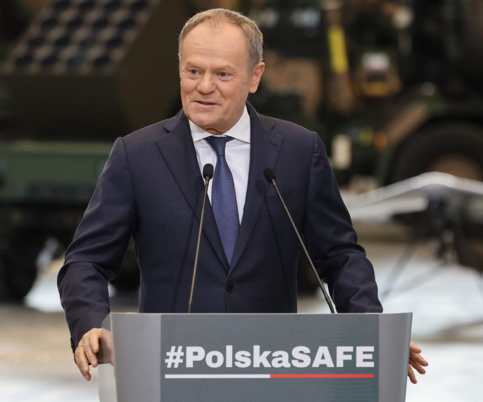 Donald Tusk, premier, podczas przemówienia za mównicą z hasłem #PolskaSAFE na tle sprzętu wojskowego. Odprężony, z uśmiechem spogląda w prawo. Na portalu Super Biznes przeczytasz o umowie SAFE i środkach dla polskiej obronności.