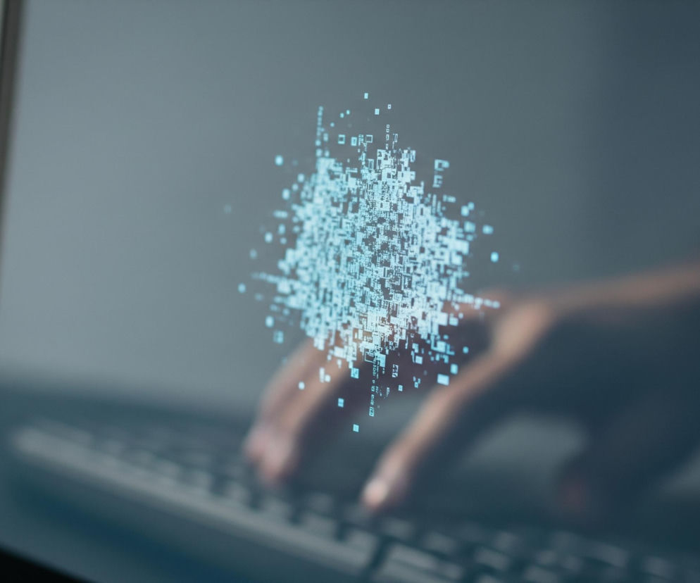 Na pierwszym planie widoczny jest monitor laptopa z częściowo widoczną klawiaturą, lekko pochylony w prawo. Ekran monitora jest ciemnoszary, a na nim widnieje jaskrawoniebieska, rozmyta plama składająca się z drobnych kształtów przypominających piksele, liter i cyfr, tworząca abstrakcyjny wzór w centrum. W tle, za ekranem, rozmyta jest dłoń osoby, która prawdopodobnie pisze na klawiaturze; widoczne są kontury palców i fragment klawiszy. Kolory w tle są również ciemne, dominują odcienie szarości i błękitu.