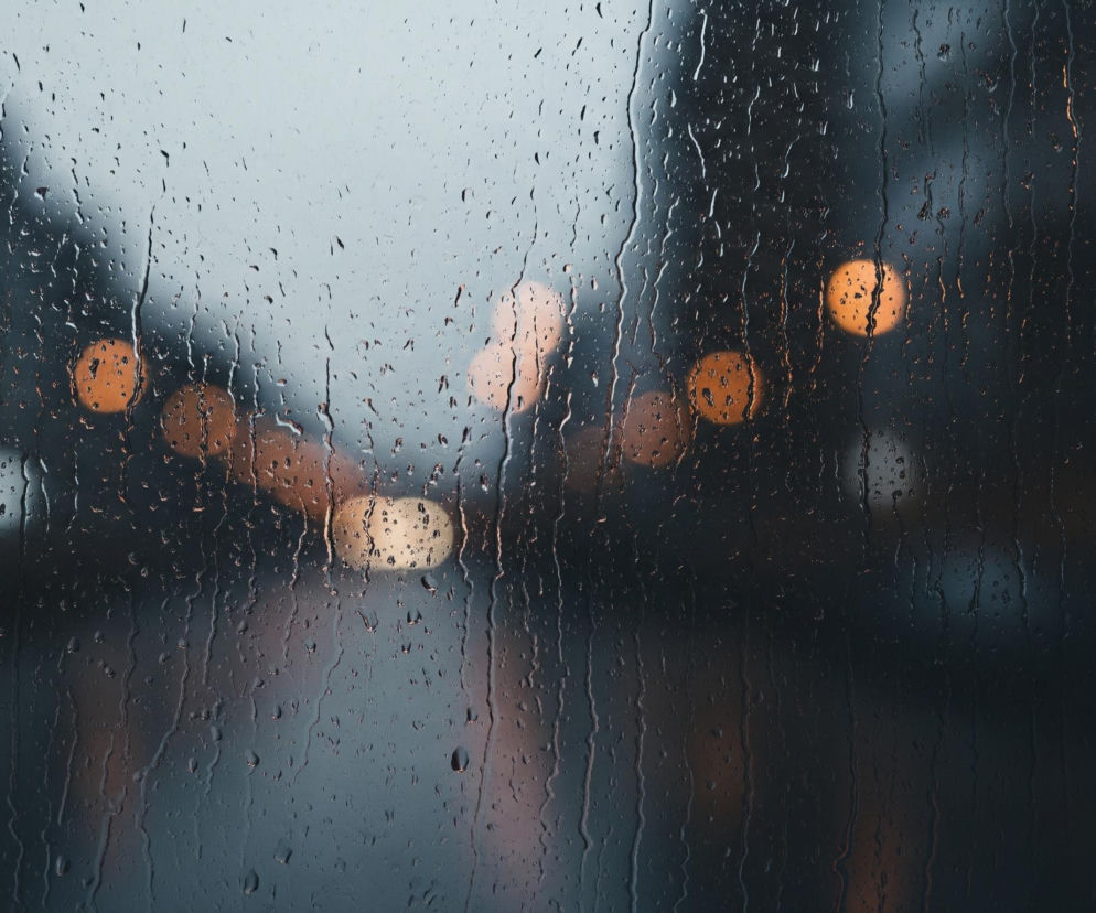 Szyba pokryta jest licznymi kroplami deszczu o różnym rozmiarze, niektóre spływają w dół, tworząc smugi. Za szybą widoczna jest rozmyta scena w ciemnych, szaro-niebieskich barwach. W tle migoczą ciepłe, pomarańczowo-żółte, rozmyte światła, przypominające lampy uliczne lub światła pojazdów. Cały obraz utrzymany jest w spokojnej, nastrojowej tonacji.