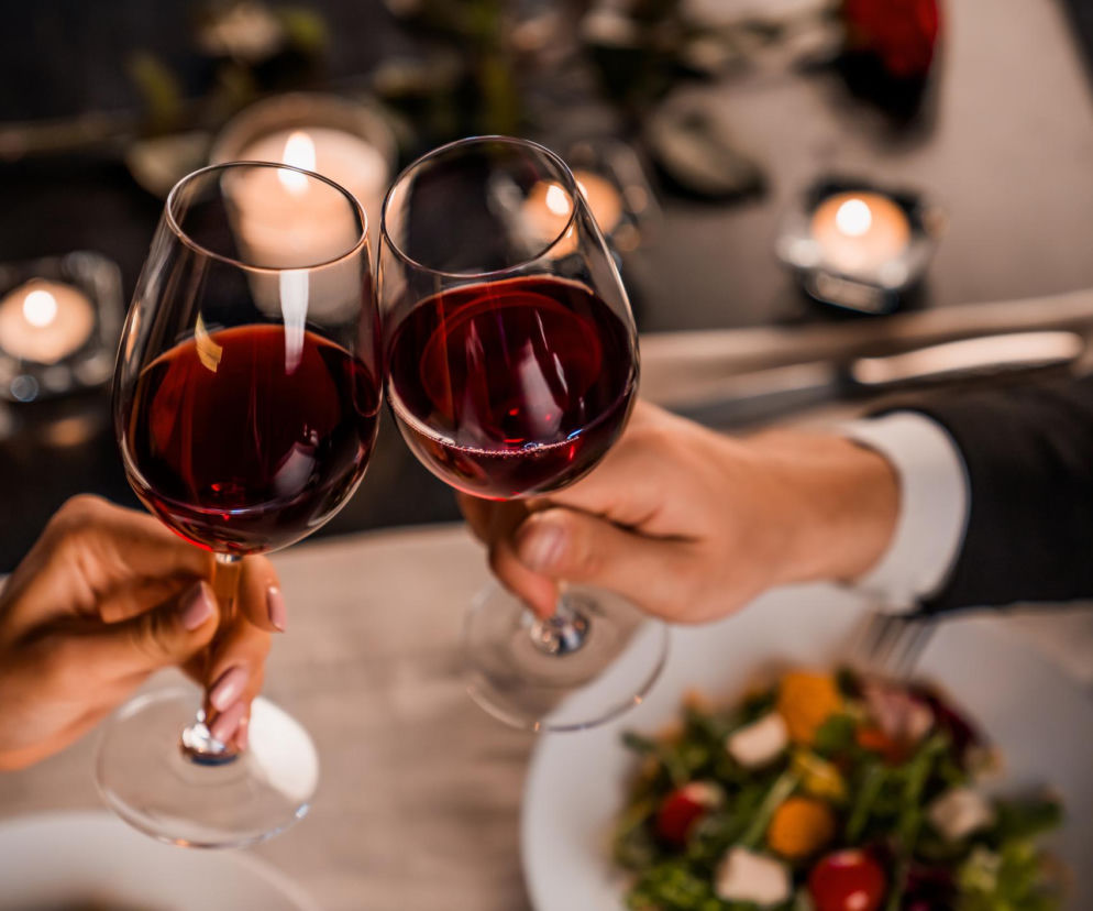 Dwie osoby wznoszące toast kieliszkami czerwonego wina podczas romantycznej kolacji przy świecach, z sałatką na stole. Artykuł na Poradnik Zdrowie wyjaśnia, dlaczego łączenie alkoholu z lekami i suplementami może być niebezpieczne.