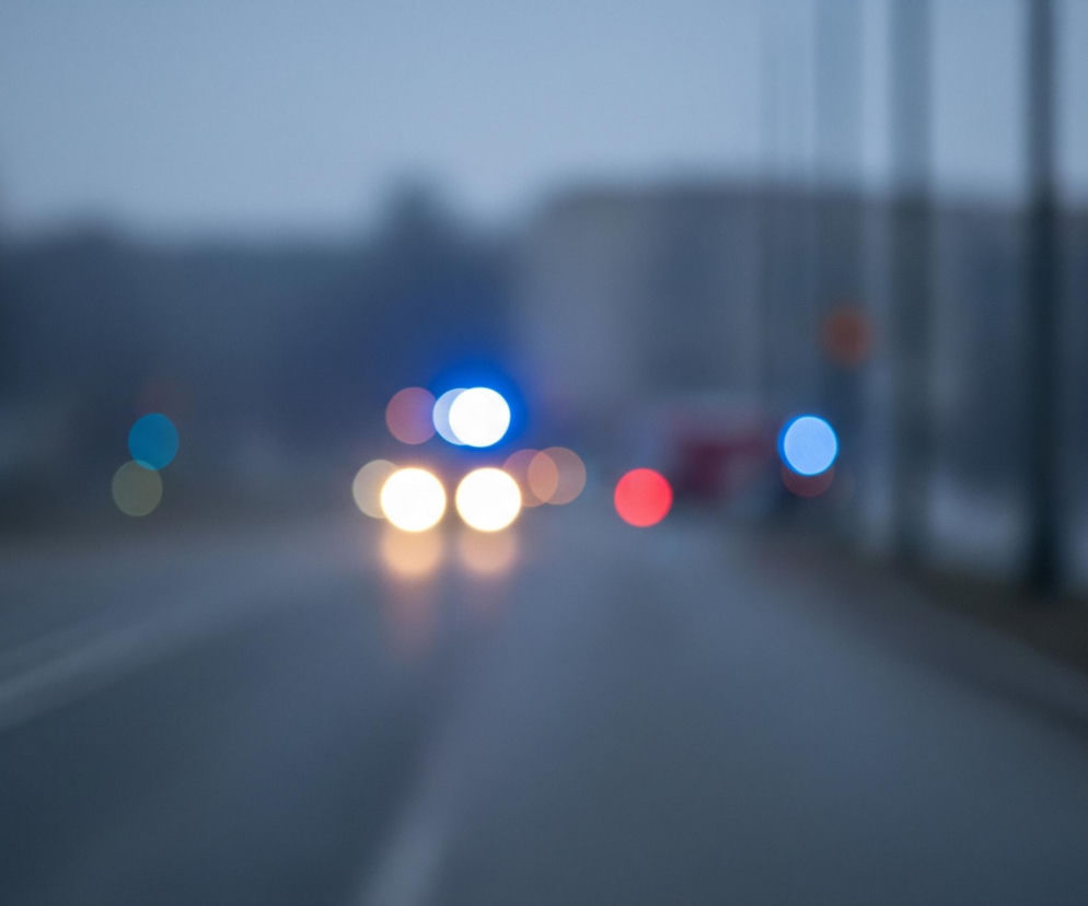 Przedstawiony obraz to rozmyte ujęcie drogi w godzinach wieczornych lub wczesnym rankiem. Na pierwszym planie, w dolnej części kadru, widoczna jest szara asfaltowa jezdnia z dwiema białymi, rozmytymi liniami oddzielającymi pasy ruchu, biegnącymi w kierunku horyzontu. W tle, na horyzoncie, znajdują się rozmyte kształty drzew i budynków, a także kilka jasnych, okrągłych świateł bokeh w różnych kolorach.

Centralne miejsce zajmują dwa wyraźne, okrągłe, biało-żółte światła samochodowe, które prawdopodobnie należą do nadjeżdżającego pojazdu. Nad nimi widoczne są dwa okrągłe, niebieskie światła, z czego jedno jest bardzo jasne i wyraźne, co sugeruje obecność pojazdu uprzywilejowanego. Po prawej stronie kadru znajdują się dodatkowe rozmyte światła, w tym jedno czerwone i jedno niebieskie. Cały obraz utrzymany jest w odcieniach szarości i błękitu, co nadaje mu zimny i tajemniczy nastrój.