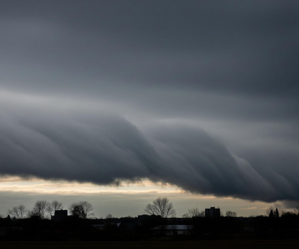 Ogromne, ciemnoszare chmury o pofalowanej, warstwowej strukturze dominują niebo. Pod spodem, wąski pas jaśniejszego, żółtawo-szarego nieba przecina horyzont. Na dole zdjęcia widać ciemną sylwetkę drzew, budynków i płaskiego terenu, tworzącą kontrastowy pas z jasno oświetlonym fragmentem nieba.