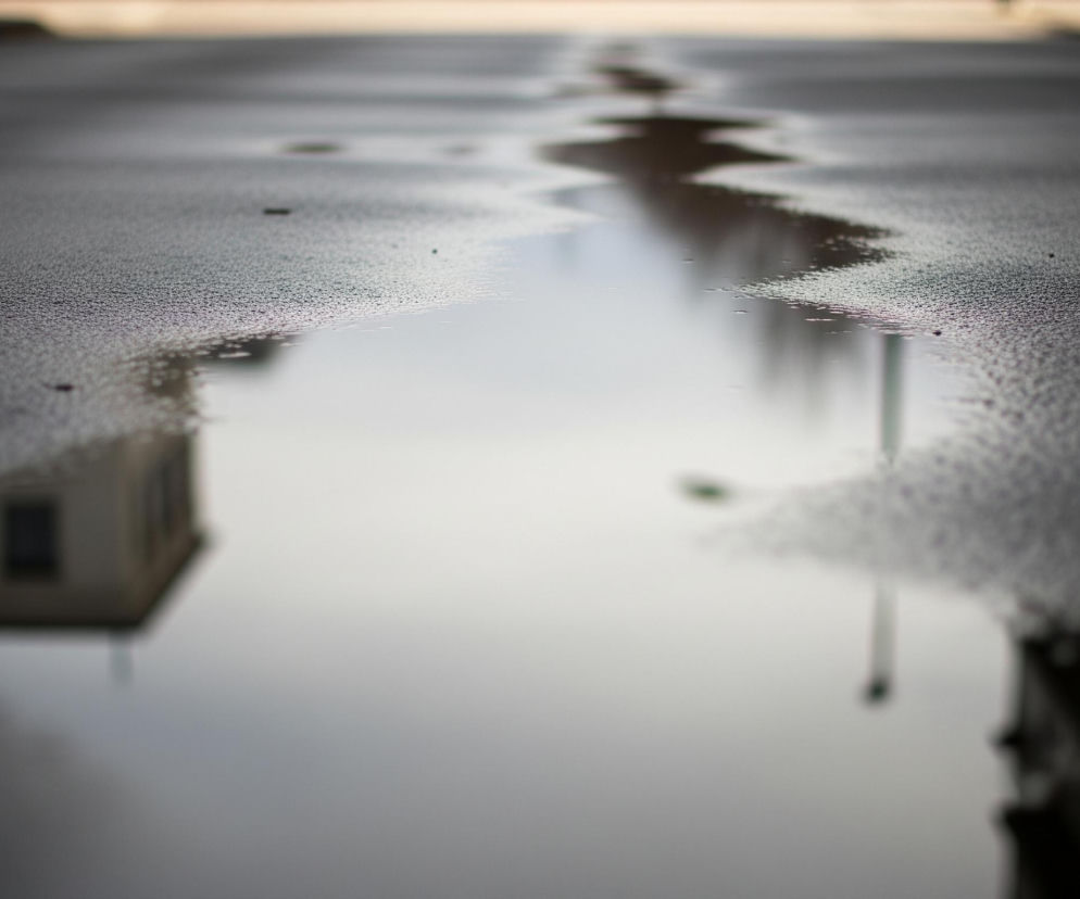 Wąski strumień wody rozciąga się wzdłuż ciemnego asfaltu, odbijając otoczenie. W lewej dolnej części odbija się jasny budynek z ciemnymi oknami. Na powierzchni wody widać również odbicie słupa z małym, zielonym elementem. Tło jest rozmyte, z ciemnymi obszarami po bokach kadru.